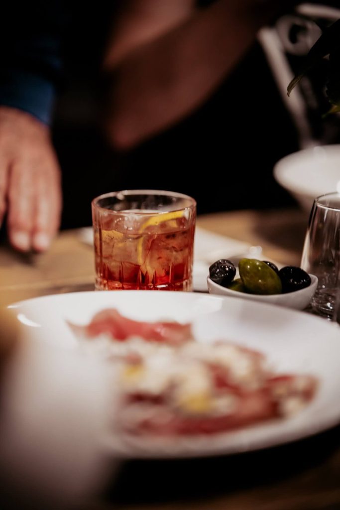 detail koktejlu v pohári od whisky, v popredí rozmazané prosciutto a olivy, v pozadí hostia