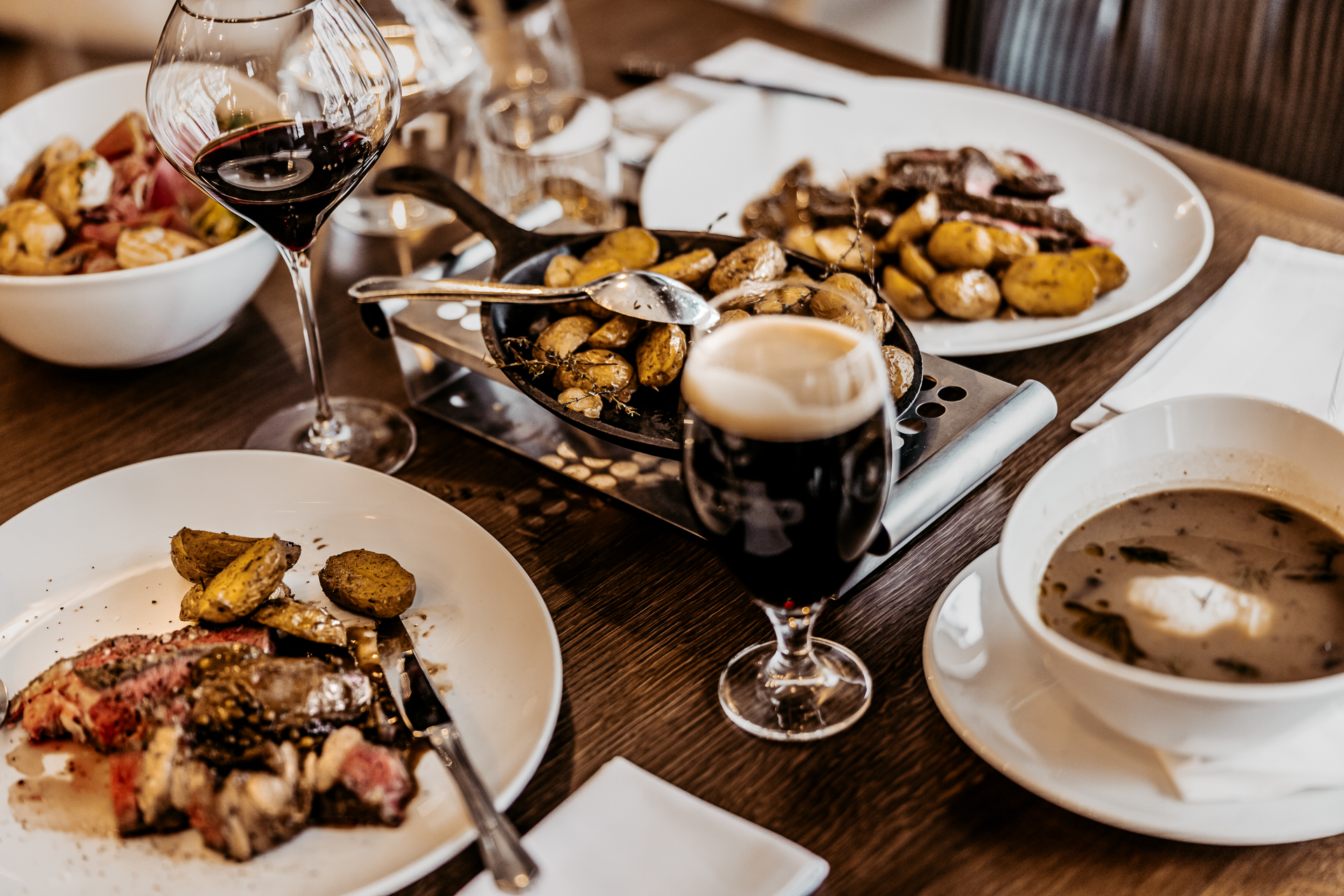 stôl plný kvalitných jedál - medium rare Tomahawk steak s opekanými zemiakmi, hríbová polievka s pošírovaným vajcom, caesar šalát, tmavé pivo a červené víno