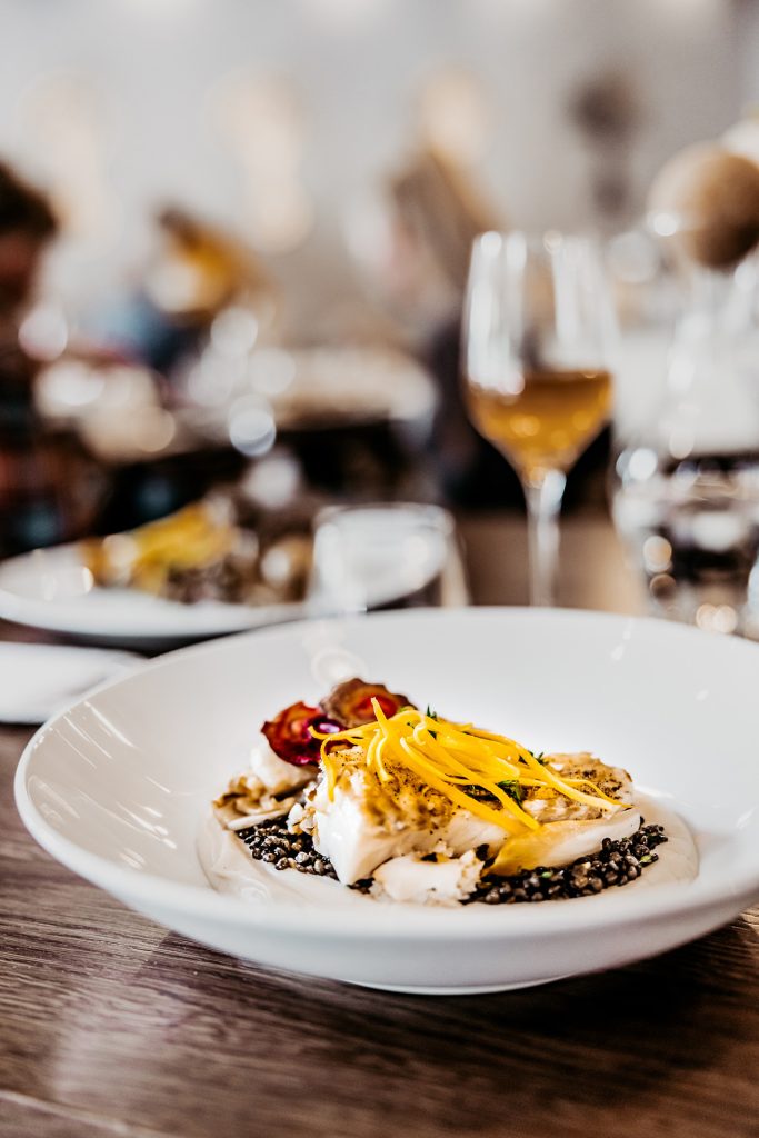 detail taniera s treskou a šošovicou, na stole s vínom, v pozadí ľudia v reštaurácii