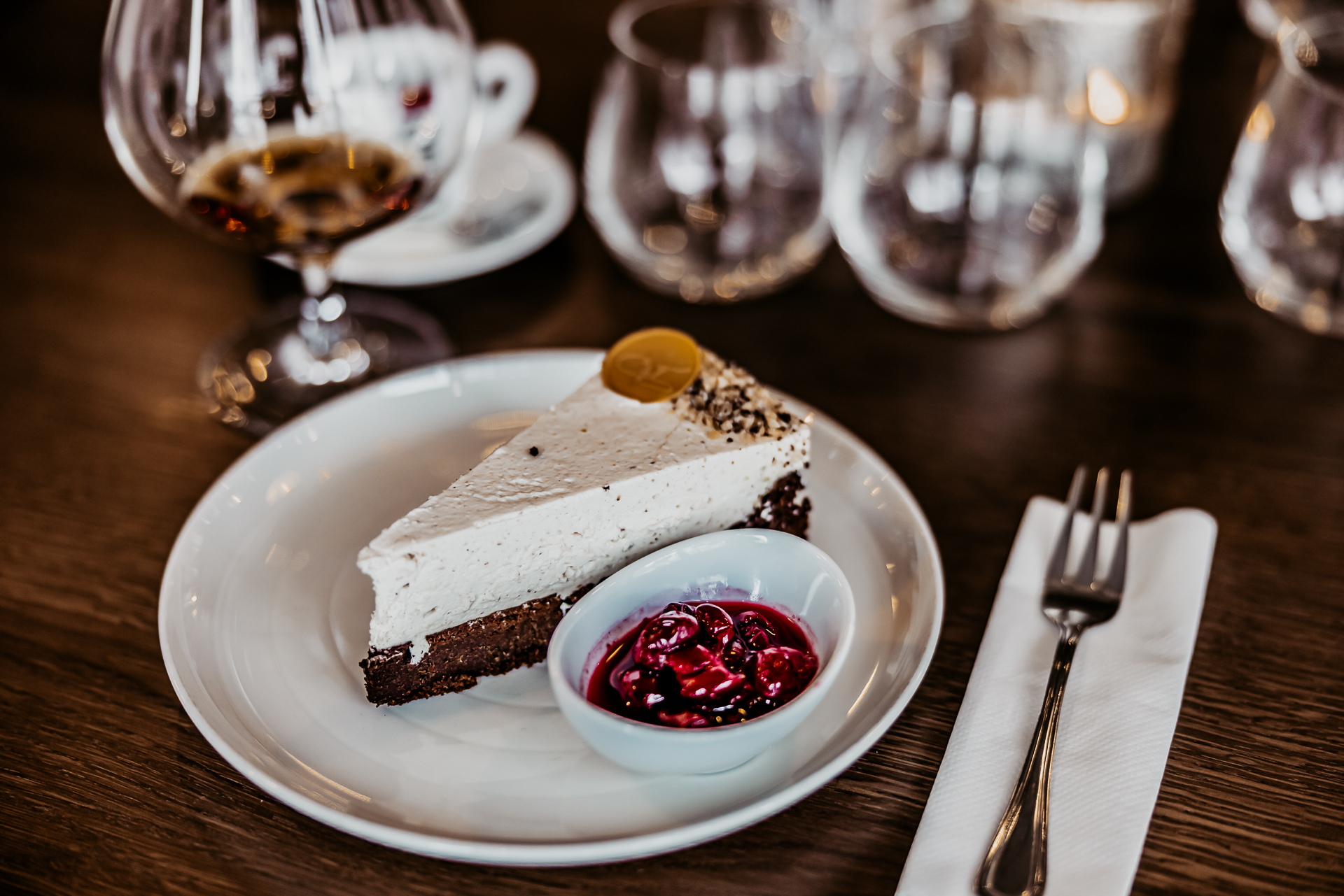 fantastická gaštanovo čokoládová torta s višňami naloženými v rume na bielom porcelánovom tanieri, na drevenom stole tiež dezertná vidlička a koňak